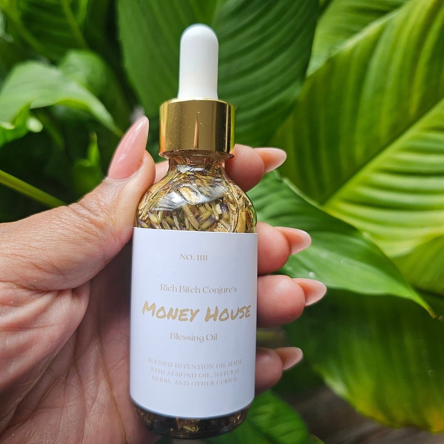 Hand holding a bottle of 'Money House' oil with green leaves in the background. Rich Bitch Conjure's Money House Blessing Oil