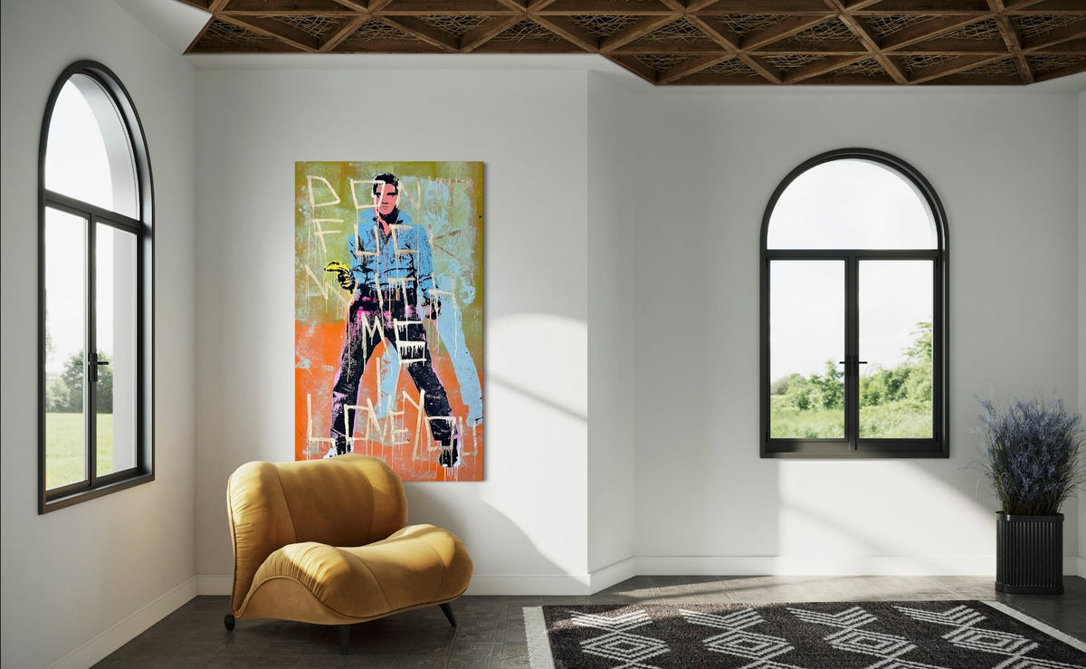 A vertical canvas art print featuring a stylized graphic illustration of a man in a blue denim jacket and black pants, displayed on a white wall in a modern room setting with a yellow armchair and arched windows. Don't Fuck With Me I Love You 2 - Silk Screen, Acrylic & Spray Paint on canvas, 2024