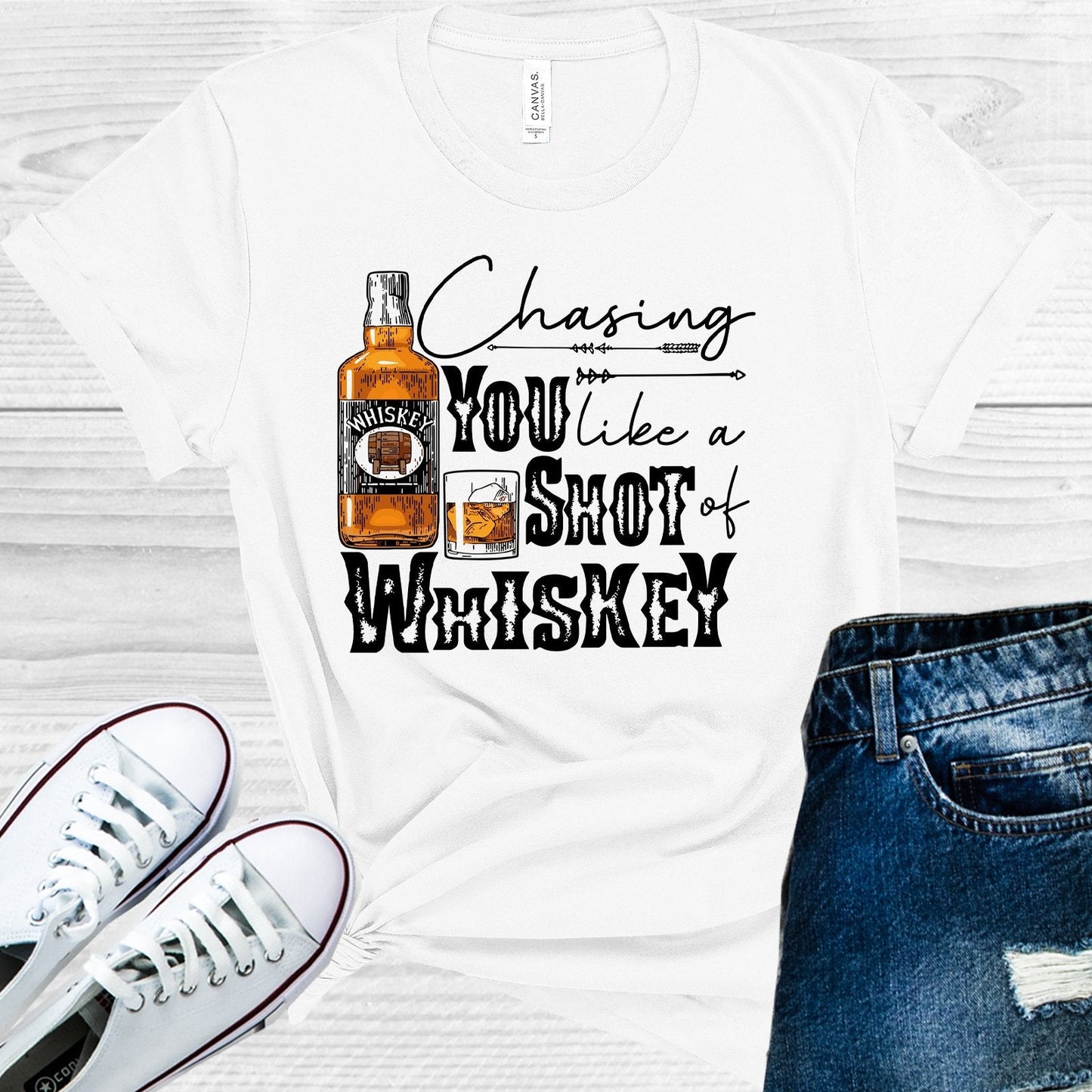 White t-shirt with whiskey graphic and text 'Chasing You Like a Shot of Whiskey' on a wooden surface with white sneakers and blue jeans.