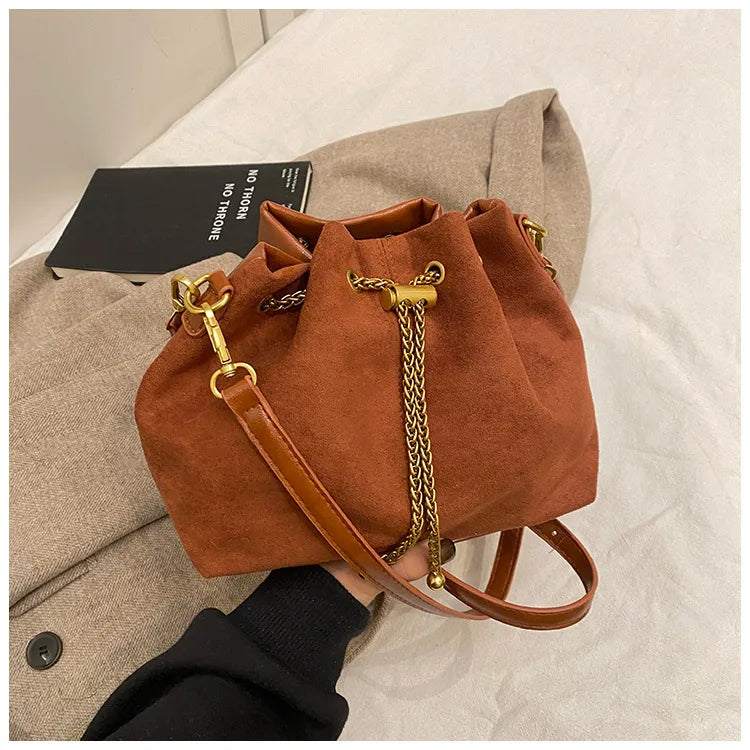 Brown frosted bucket handbag with gold chain drawstring closure and adjustable shoulder strap on a beige fabric background. Stylish women's PU leather crossbody purse with a retro-inspired design for everyday use.