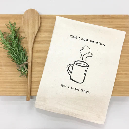 Napkin with coffee-themed text and graphic on a wooden cutting board with a spoon and rosemary. Kitchen towel that reads First I drink coffee then I do the things.