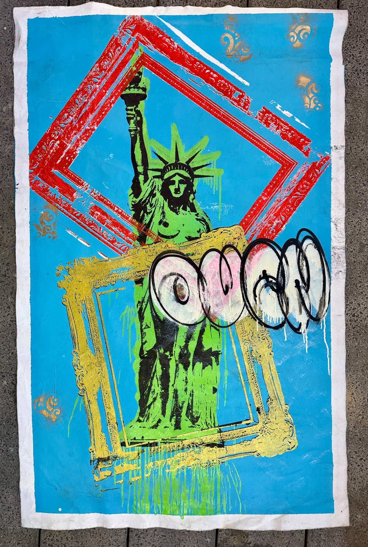 A square textile or poster-like product laid flat on a gray textured surface, featuring a bright blue field with a green Statue of Liberty graphic, red and yellow frame-like brushstroke elements, and stylized graffiti lettering across the center. Framed Liberty