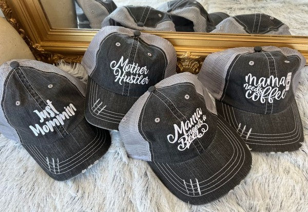 Vintage charcoal gray trucker hats embroidered with funny mom sayings including "Mama Needs a Drink," "Mama Needs Coffee," "Mother Hustler," and "Just Mommin'," displayed on a soft faux fur surface with a decorative mirror in the background. Mama Needs a Drink Embroidered Trucker Hat – Funny Mom Mesh Baseball Cap | Adjustable Snapback Hat for Mothers