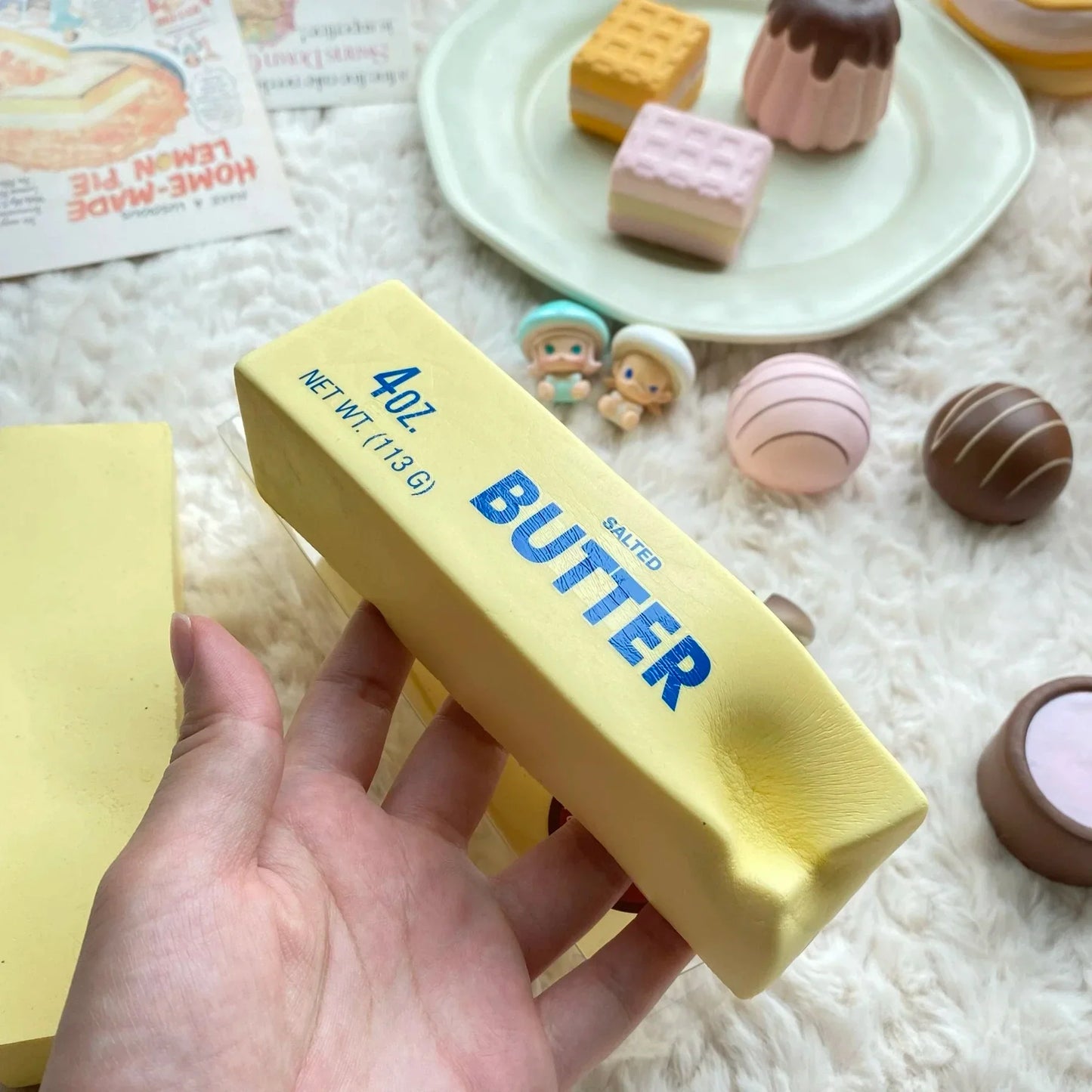 Hand holding a realistic yellow butter stick squishy toy labeled "Butter" with 4 oz (113 g) packaging, surrounded by pastel dessert squishies, on a soft white plush background. Squishy toy.