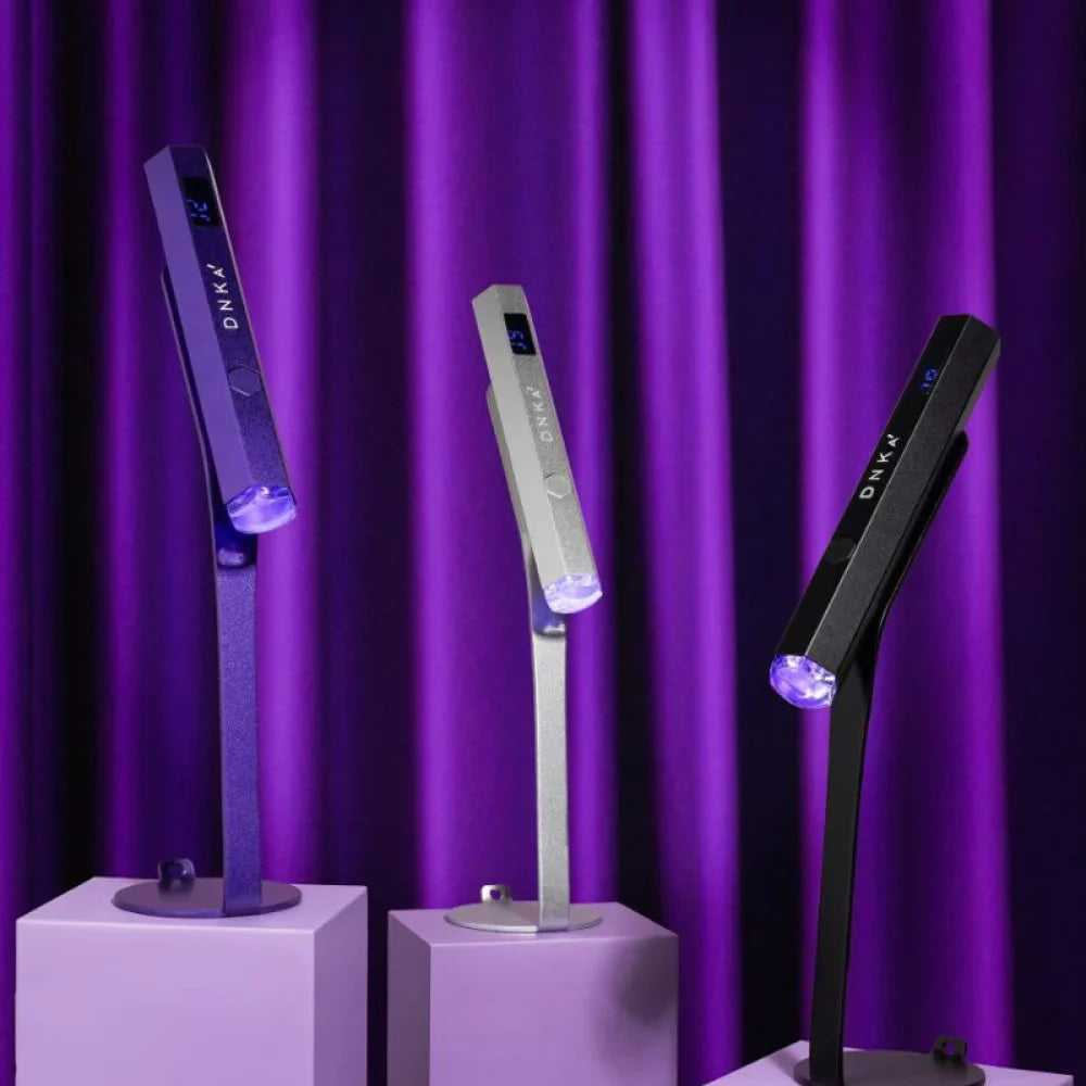Three portable LED nail lamps in black, silver, and purple, each mounted on a matching colored display stand against a purple curtain background.