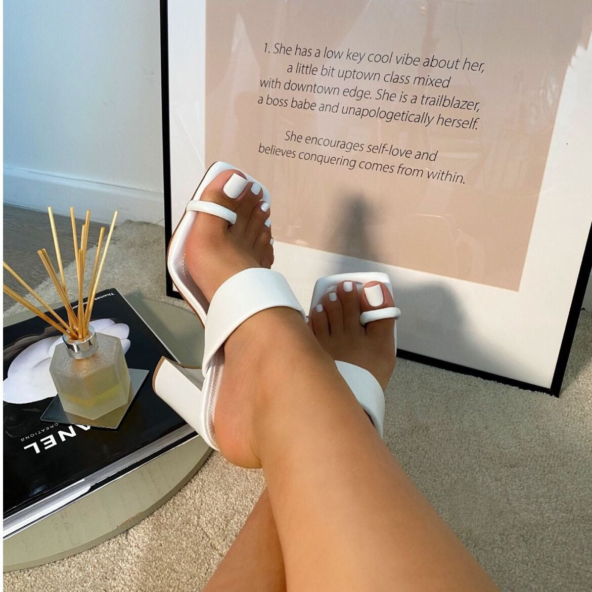 White sandals on a foot with a blurred background featuring a quote and a diffuser.