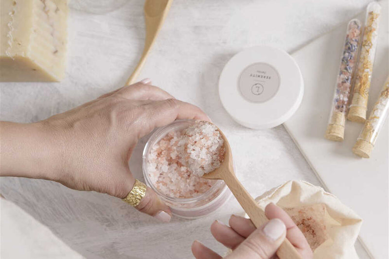 Natural Lavender Bath Salts in a glass jar alongside a wooden scoop and a small linen bag on a clean white surface.