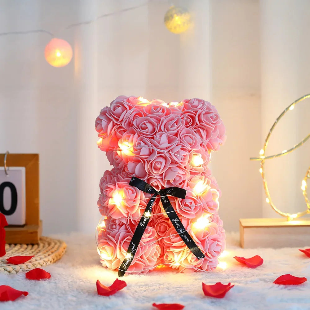 Pink rose bear with lights on a decorative surface with red petals and string lights in the background.