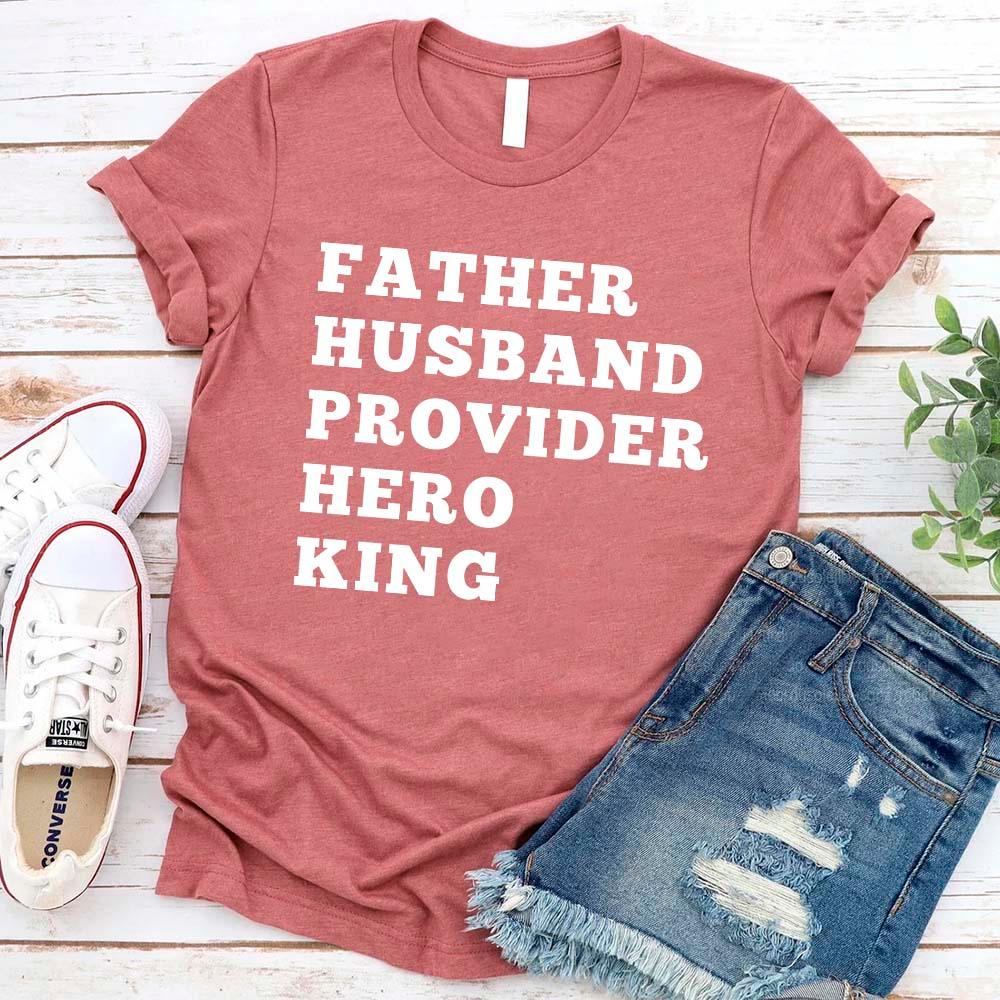 Red t-shirt with text 'FATHER HUSBAND PROVIDER HERO KING' on a wooden surface with shoes and shorts.