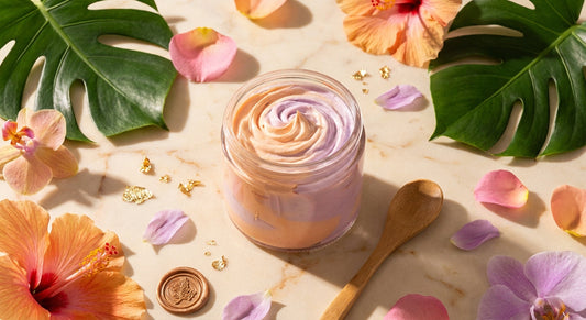 A luxurious flat lay of artisan whipped body butter in a glass jar surrounded by gold leaf flakes, colorful petals, and tropical botanicals on a warm marble surface.
