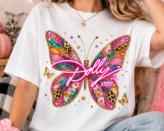 A person wearing a white short-sleeve T-shirt with a large colorful butterfly graphic and the text 'Dolly XOXO' across the front, photographed in a cozy indoor setting.