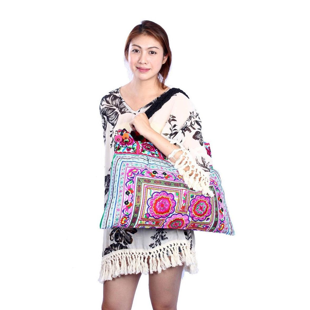 A woman smiling while holding a colorful, handcrafted boho-style tote bag featuring intricate embroidery and vibrant pom-pom fringe details, set against a clean, minimalist white background.