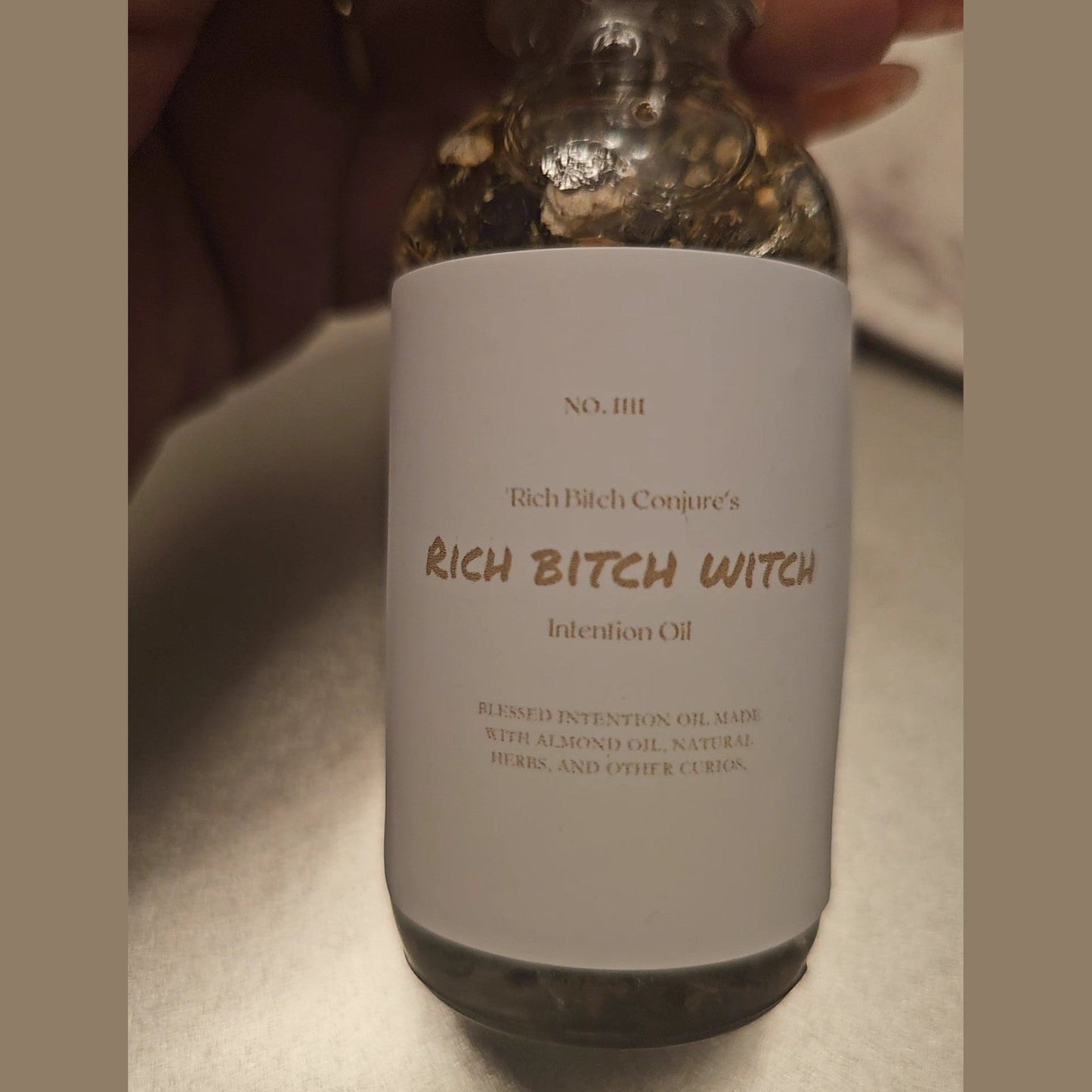Bottle labeled 'Rich Bitch Conjure's Rich Bitch Witch Intention Oil' held by a person. Rich Bitch Conjure's Rich Witch Bitch Intuitive Oil