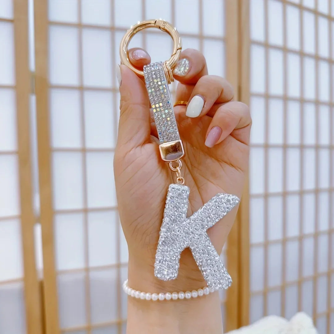 Sparkling rhinestone letter **K** keychain with a gold-tone ring and clasp, held in hand against a neutral background. Crystal-covered initial charm doubles as a stylish bag charm or key accessory.
