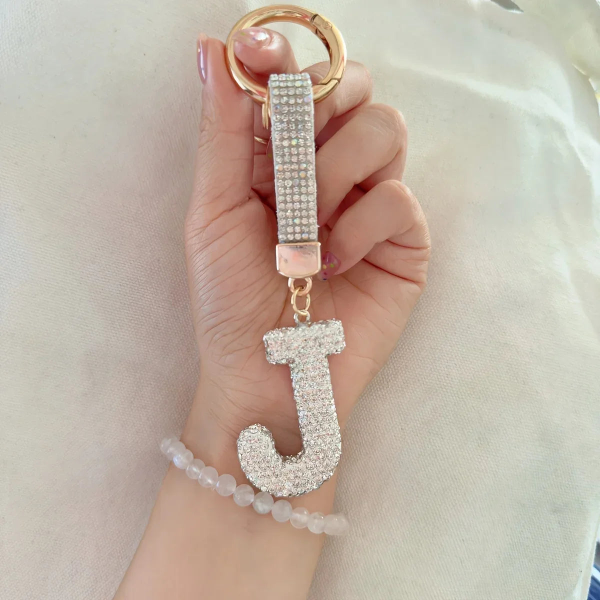 Hand holding a gold-tone rhinestone initial J keychain with a sparkling crystal letter charm and matching rhinestone wrist strap against a soft neutral fabric background. Decorative alphabet keychain with round key ring, perfect for bags, purses, backpacks, or keys.