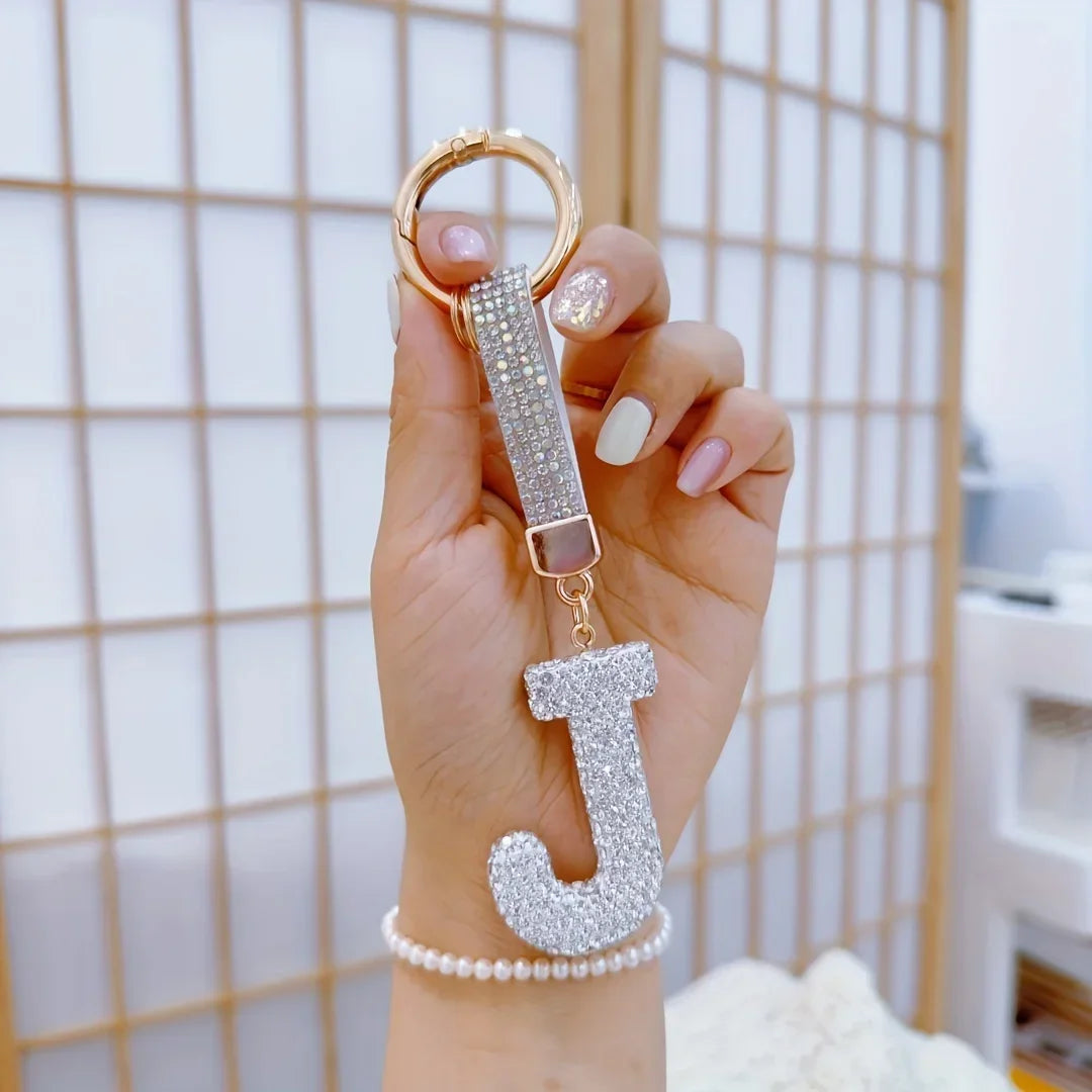 Hand holding a sparkling rhinestone letter **J** keychain with a gold-tone ring and crystal-studded strap against a soft neutral background, showcasing a personalized alphabet bag charm and keyring accessory.
