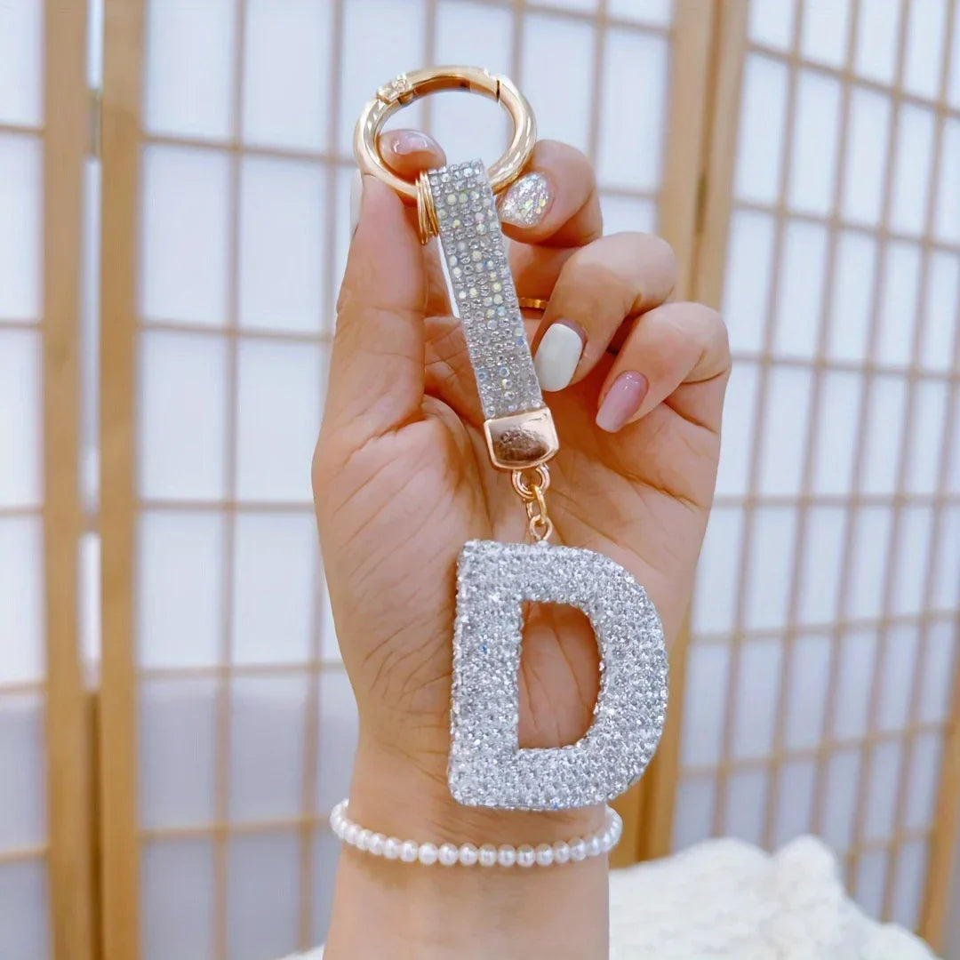 Sparkling rhinestone letter D keychain with a gold-tone ring and crystal-studded strap, held in hand against a soft neutral background. Decorative alloy initial charm for keys, handbags, backpacks, or gifts.
