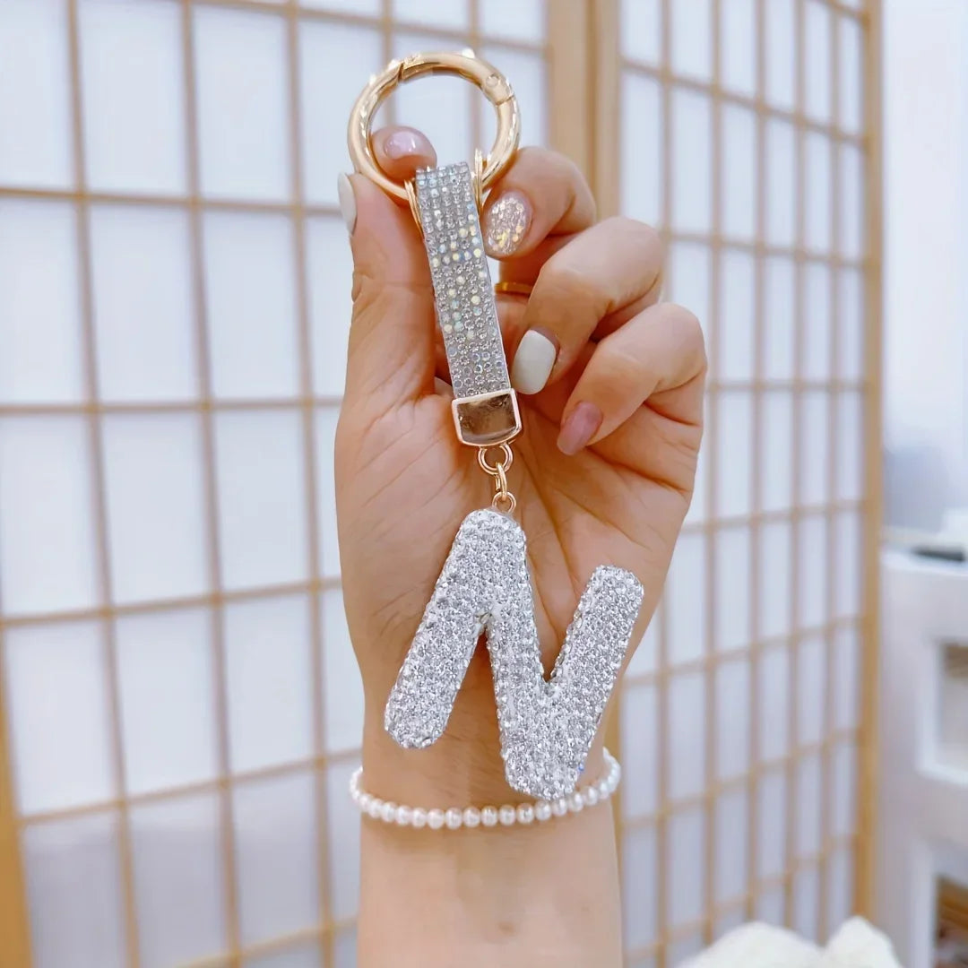 A close-up of a hand holding a gold-tone rhinestone keychain featuring a sparkling silver letter "N" pendant. The decorative alphabet charm hangs from a round key ring with a crystal-studded strap against a soft neutral background.
