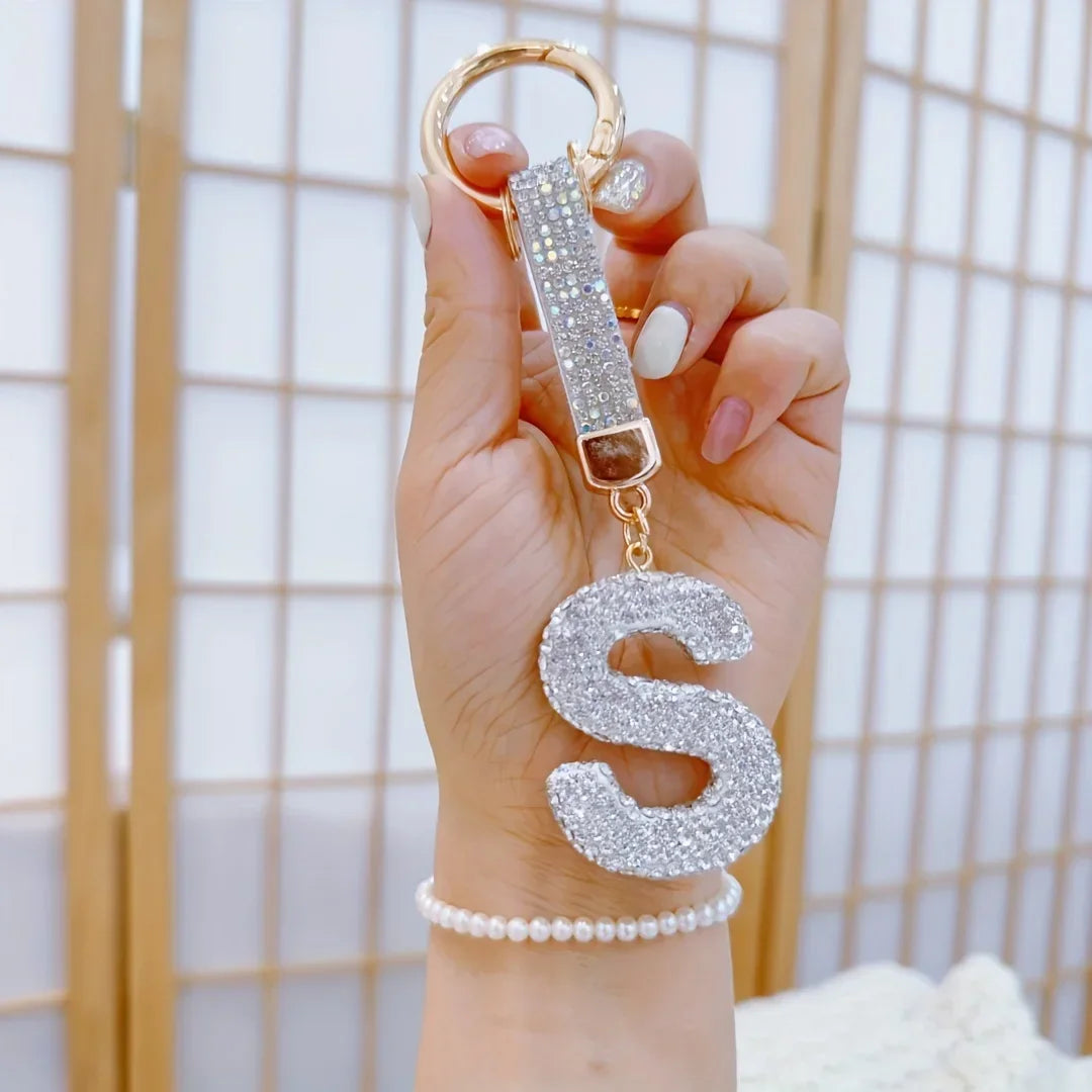 Sparkling rhinestone letter "S" keychain with a gold-tone ring and crystal-covered initial charm, held in hand against a soft neutral background. Decorative alloy alphabet keychain accessory for keys, handbags, backpacks, or gifts.