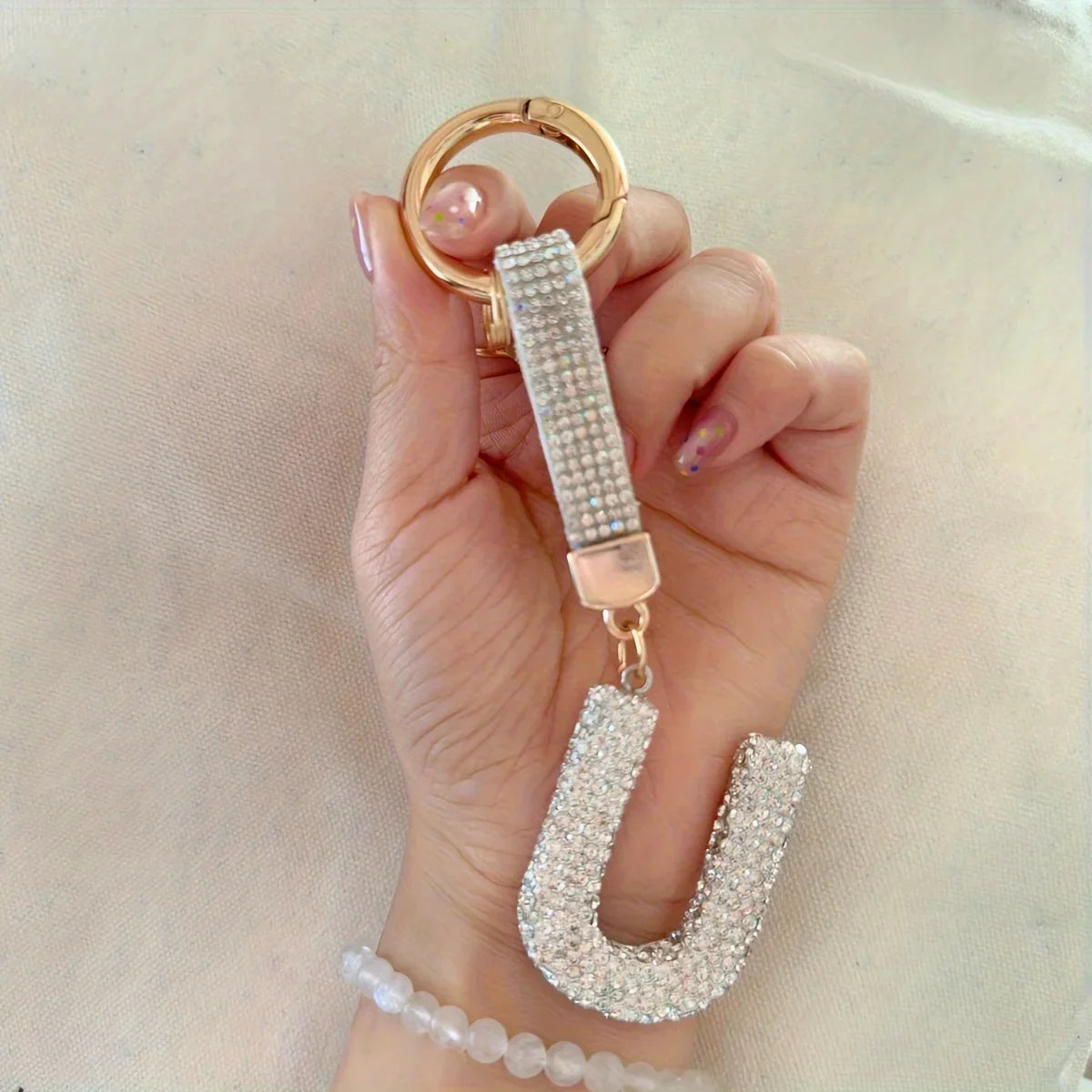 Hand holding a sparkling rhinestone alphabet keychain with a gold-tone key ring and crystal-covered letter charm against a neutral fabric background. Decorative initial keychain accessory with a beaded wrist strap, ideal for handbags, backpacks, keys, or gifts.