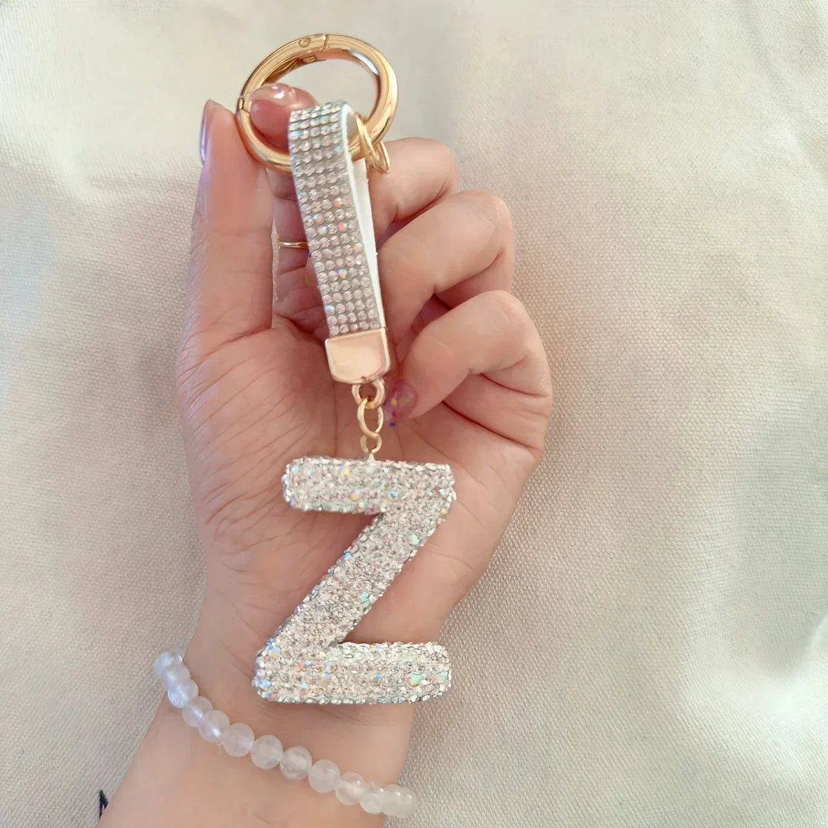 Hand holding a gold-tone rhinestone alphabet **"Z"** keychain with a matching wrist strap against a soft neutral fabric background. Sparkling crystal letter charm with round key ring, shown as a stylish handbag accessory.
