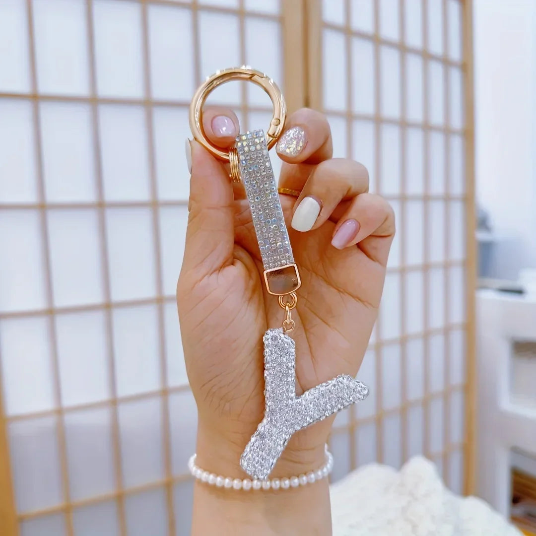 Hand holding a sparkling rhinestone letter **Y** keychain with a gold-tone ring and crystal-studded strap against a softly blurred indoor background. Decorative alphabet charm accessory for handbags, backpacks, keys, and personalized gifts.