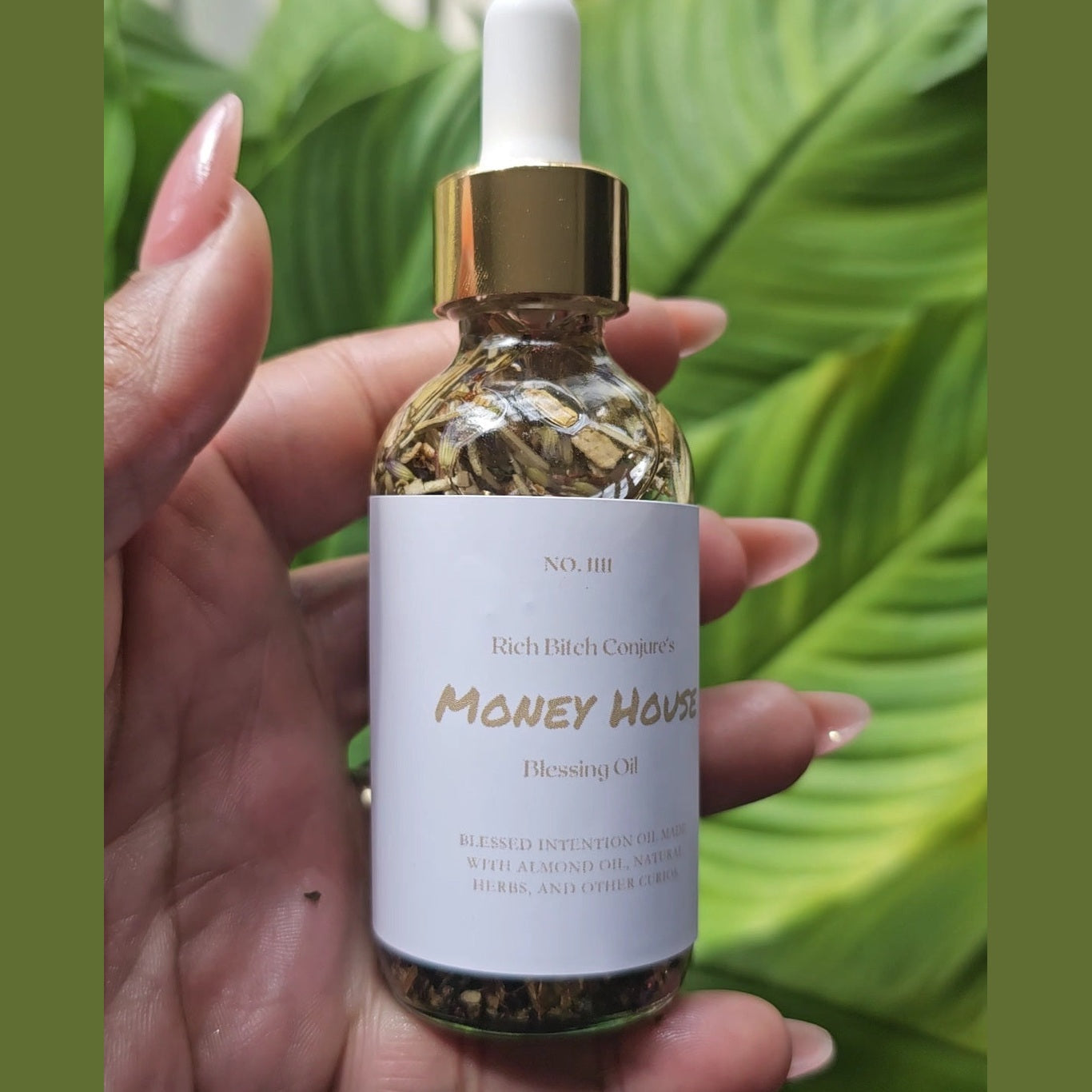 Hand holding a bottle of 'Money House' blessing oil with a gold cap against a green leafy background. Rich Bitch Conjure's Money House Blessing Oil