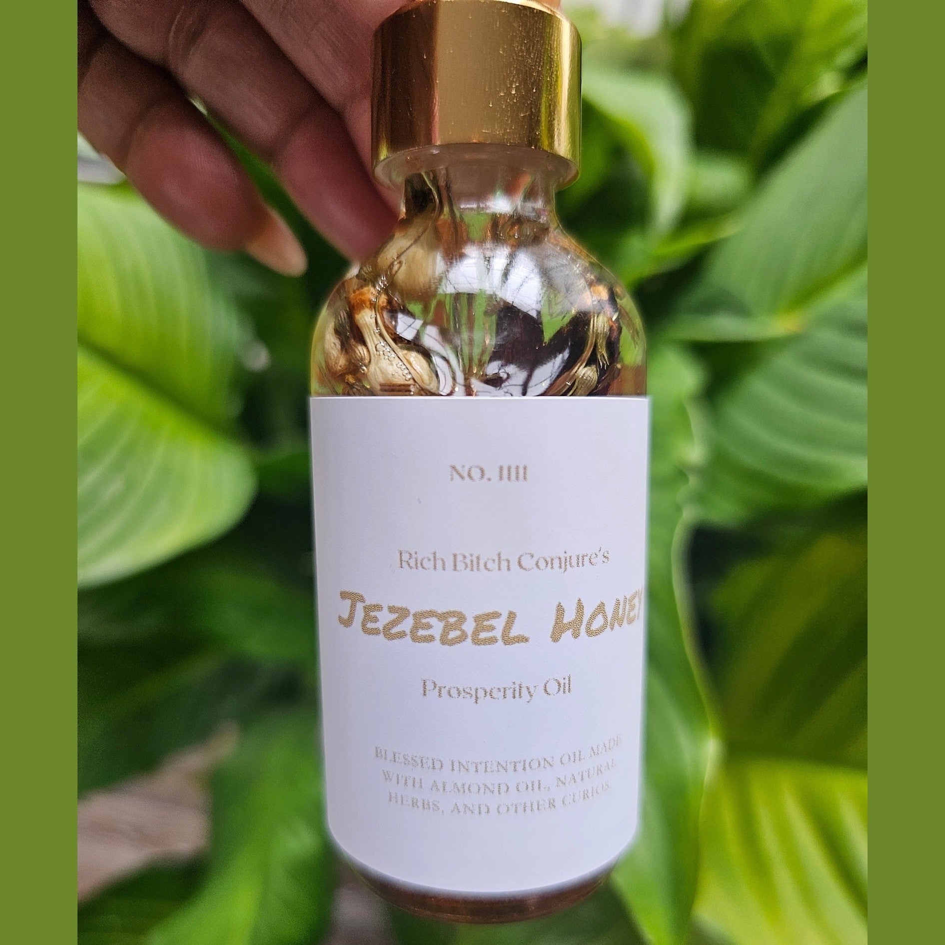 Bottle of 'Jezebel Honey' prosperity oil with a gold cap against a green leafy background. Rich Bitch Conjure's Jezebel Honey Oil