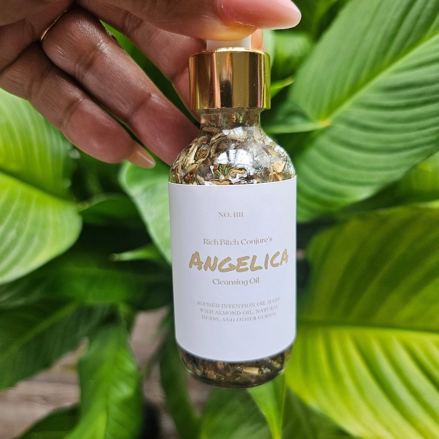 Bottle of Angelica cleansing oil held by a hand with green leaves in the background. Rich Bitch Conjure's Cleansing Oil  | Angelica