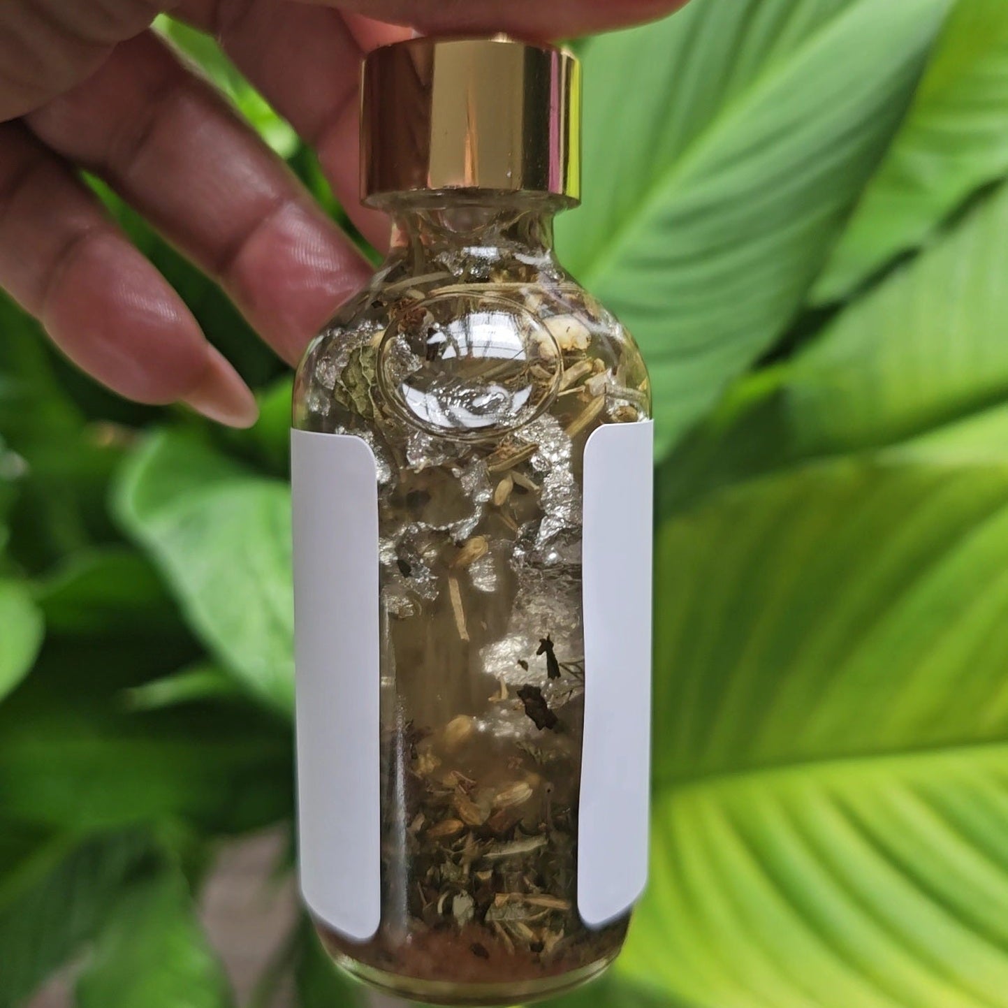 Clear bottle with gold cap and white label held by a hand against a green leafy background. Rich Bitch Conjure's Can't Touch This Protection Oil