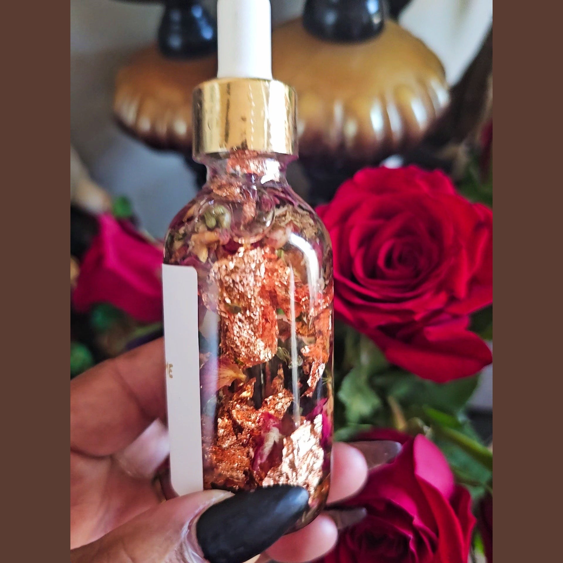 Bottle with gold cap and rose petals held in front of red roses. Rich Bitch Conjure's Come To Me Attraction Oil