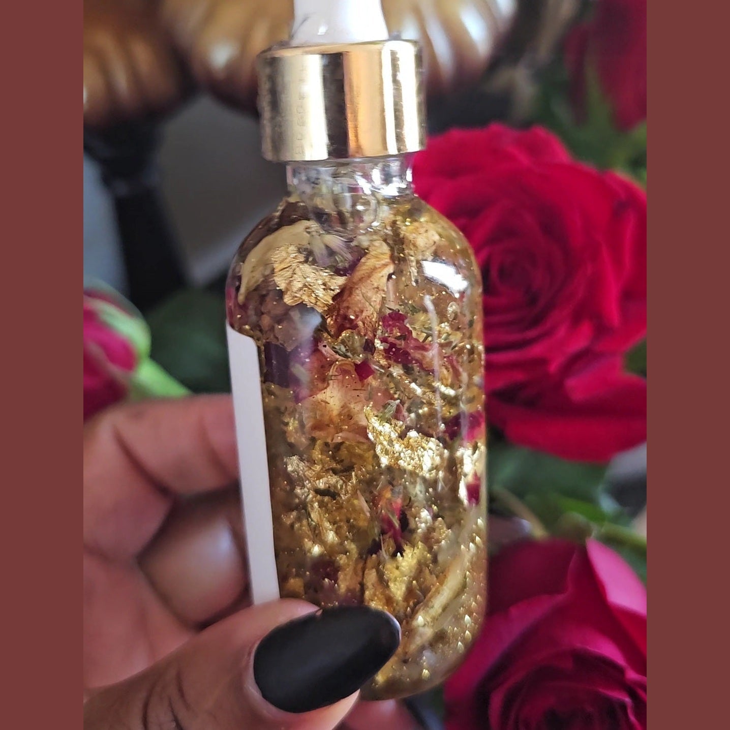 Glass bottle with gold flakes and a dropper, held in front of red roses. Lucky Rich Girl Hoodoo Conjure Oil | Wealth, Beauty, Glamour & Good Fortune Conjure Oil | Limited Edition