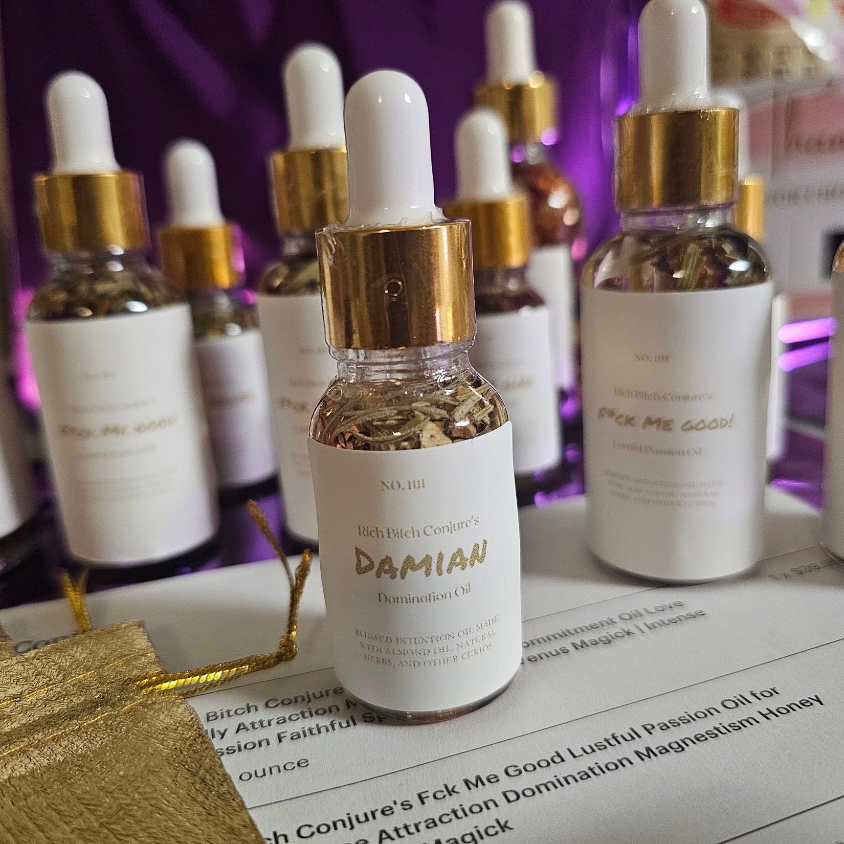 Bottle of 'Damian' oil with dropper on a table with other similar bottles in the background. Rich Bitch Conjure's Damian Oil for Intense Domination