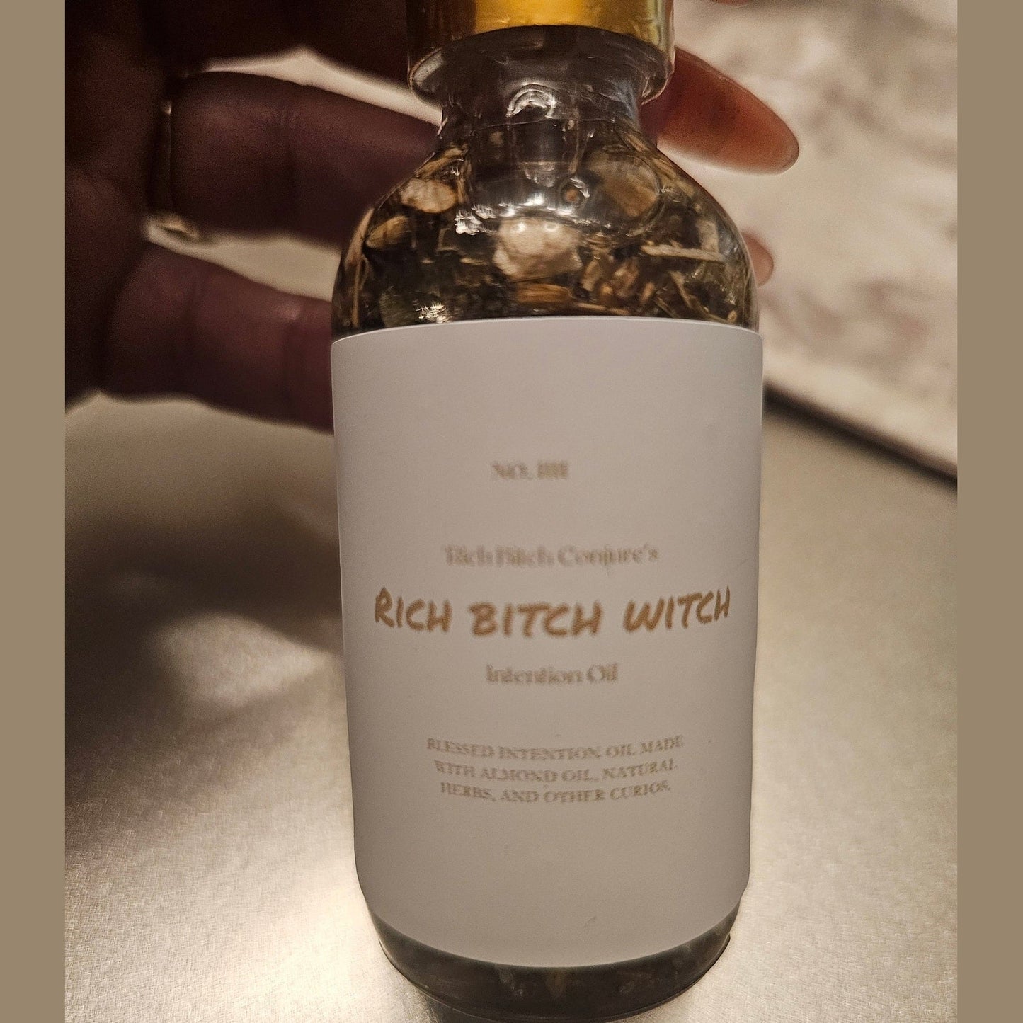 Bottle labeled 'Rich Bitch Witch' with a gold cap held by a hand. Rich Bitch Conjure's Rich Witch Bitch Intuitive Oil