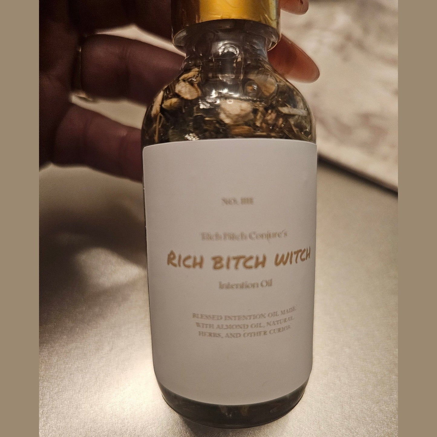 Bottle labeled 'Rich Bitch Witch' held by a hand on a neutral background. Rich Bitch Conjure's Rich Witch Bitch Intuitive Oil
