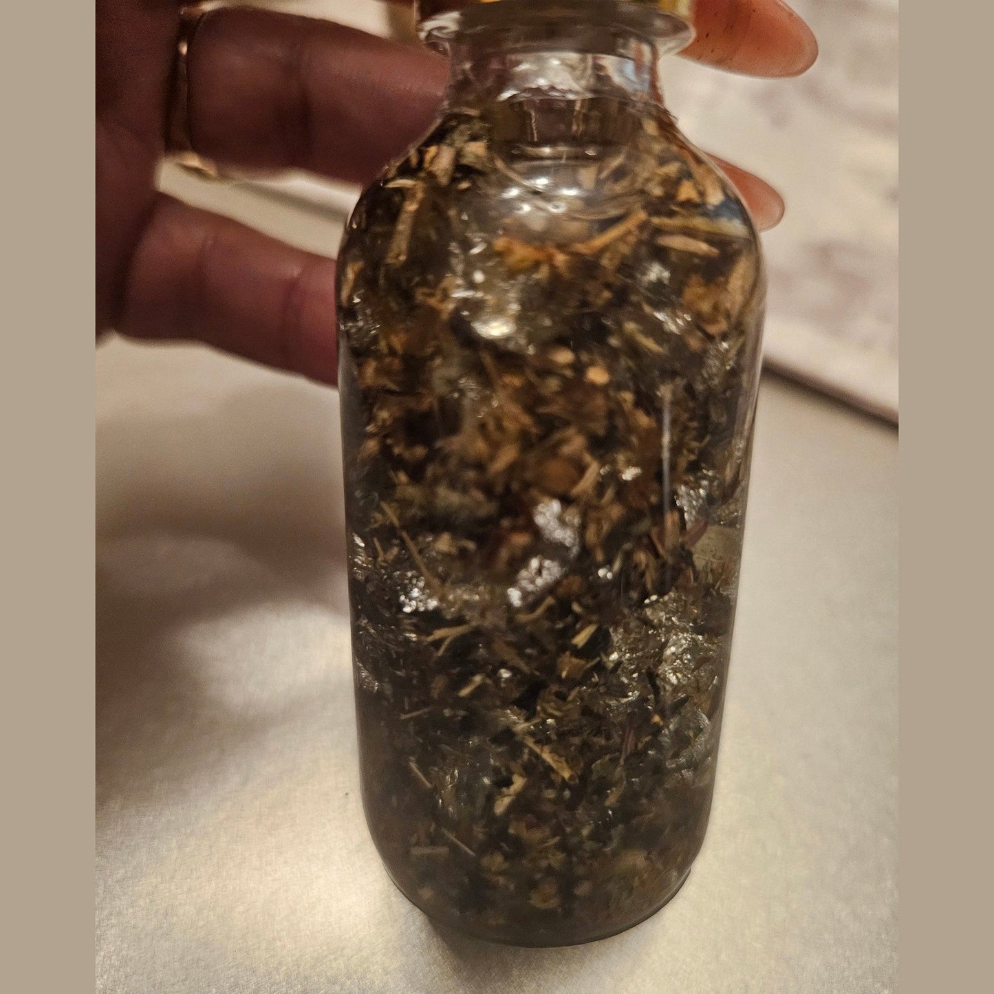 Clear glass bottle filled with dried herbs or spices held by a hand on a light background. Rich Bitch Conjure's Rich Witch Bitch Intuitive Oil