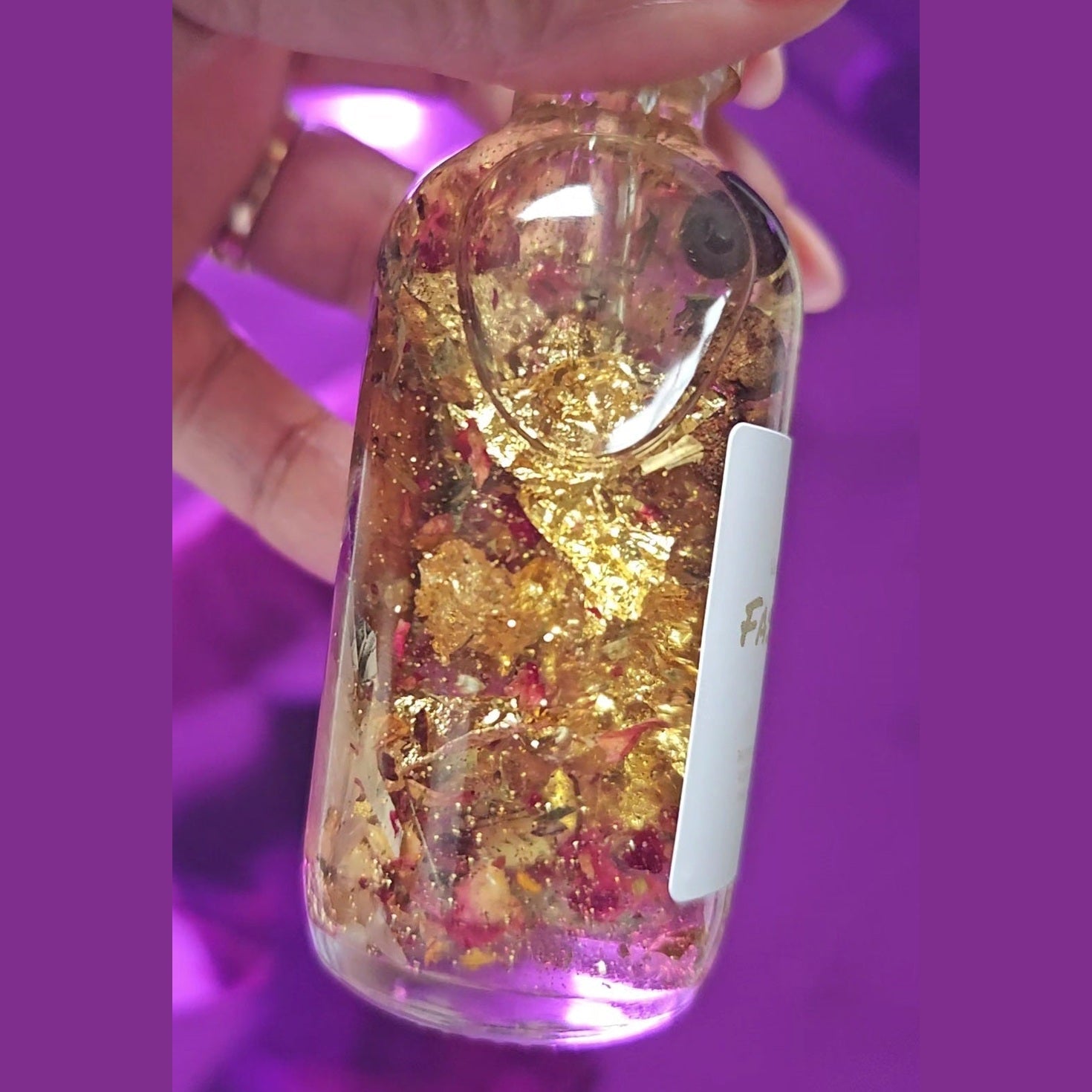 Clear bottle with gold and red contents held by a hand against a purple background. Rich Bitch Conjure's Fast Luck Money Oil