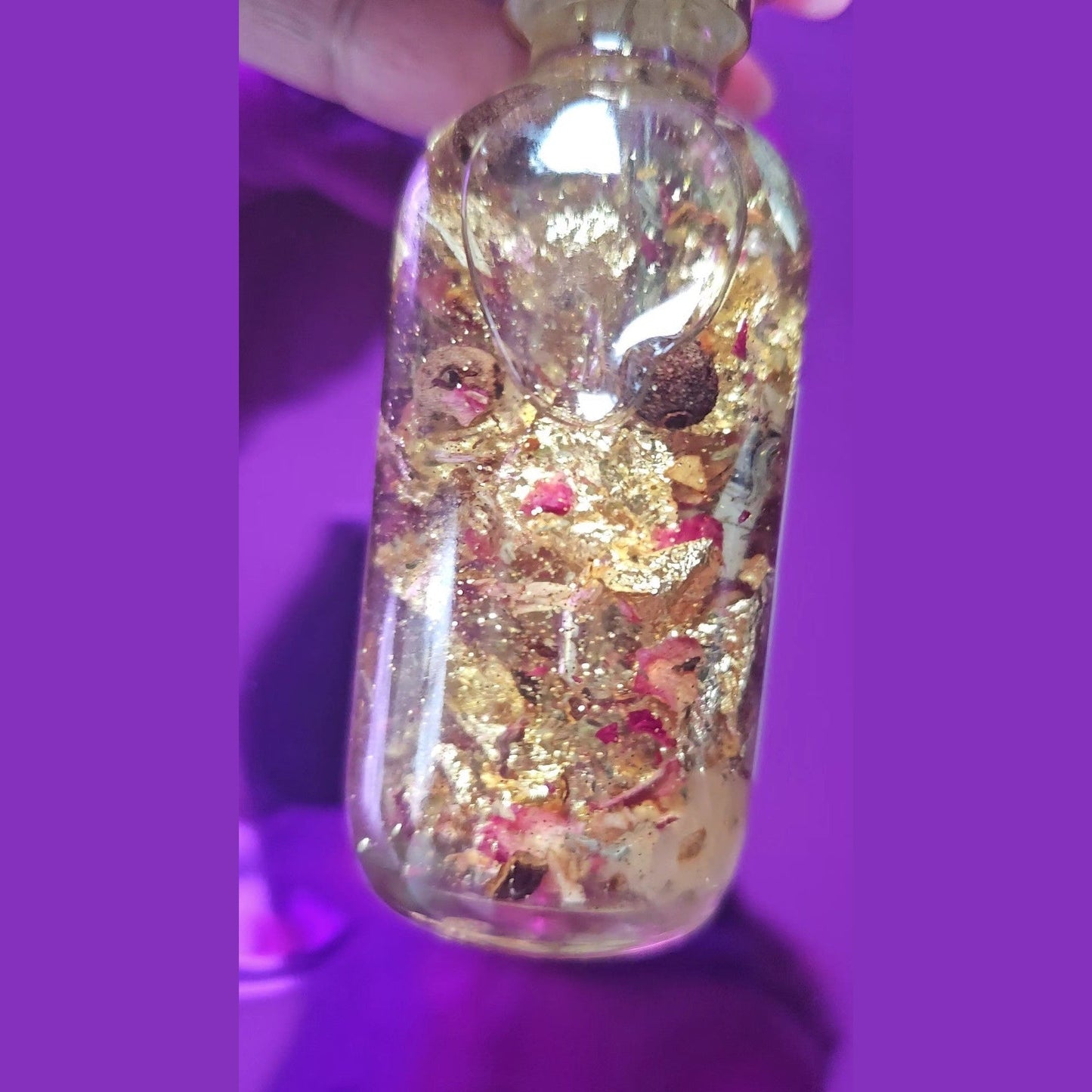 Glass bottle with gold and pink contents against a purple background. Rich Bitch Conjure's Fast Luck Money Oil