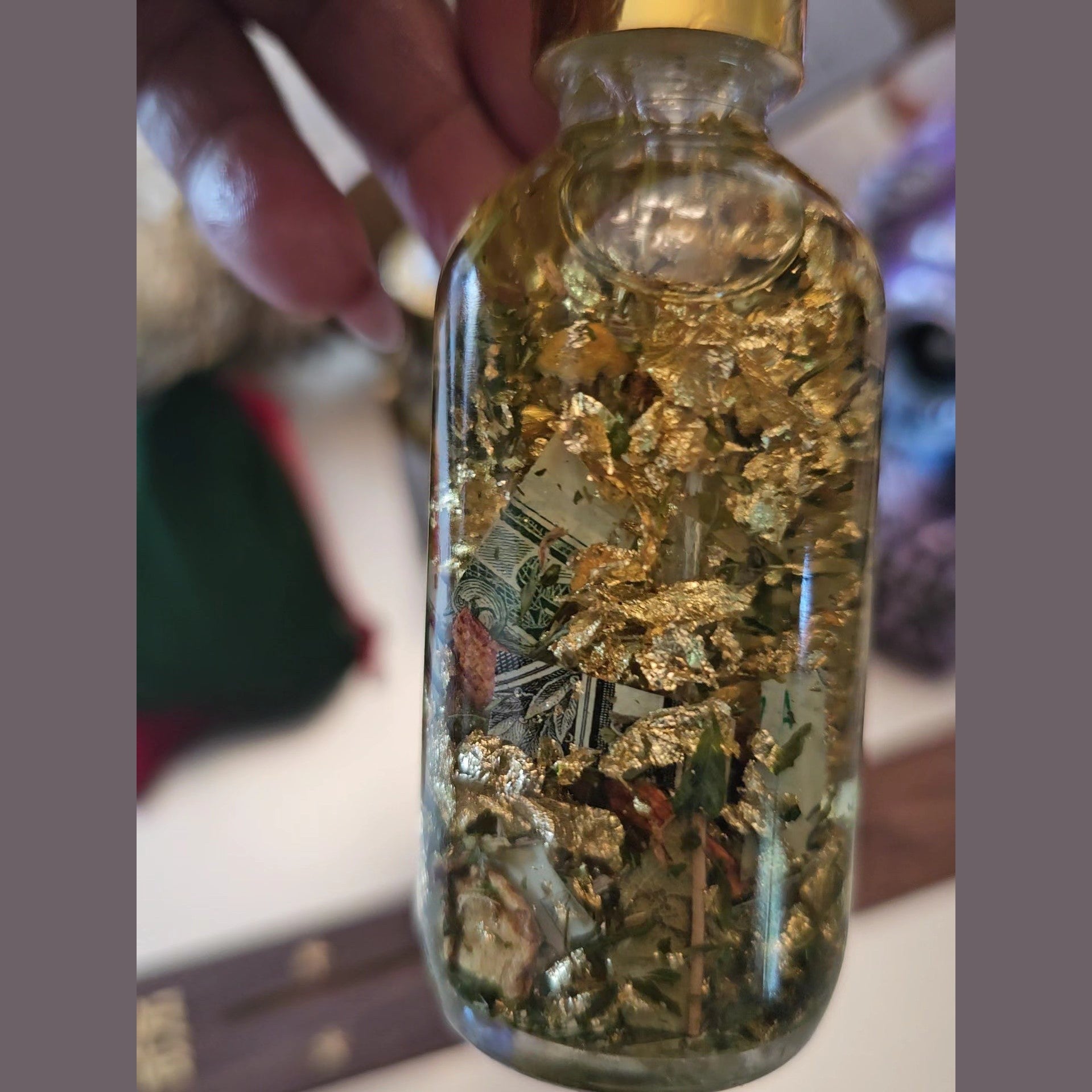 Glass bottle filled with gold leaf and other materials, held by a hand. Fast Money Oil - Luxury Hoodoo Conjure Oil for Quick Cash & Prosperity