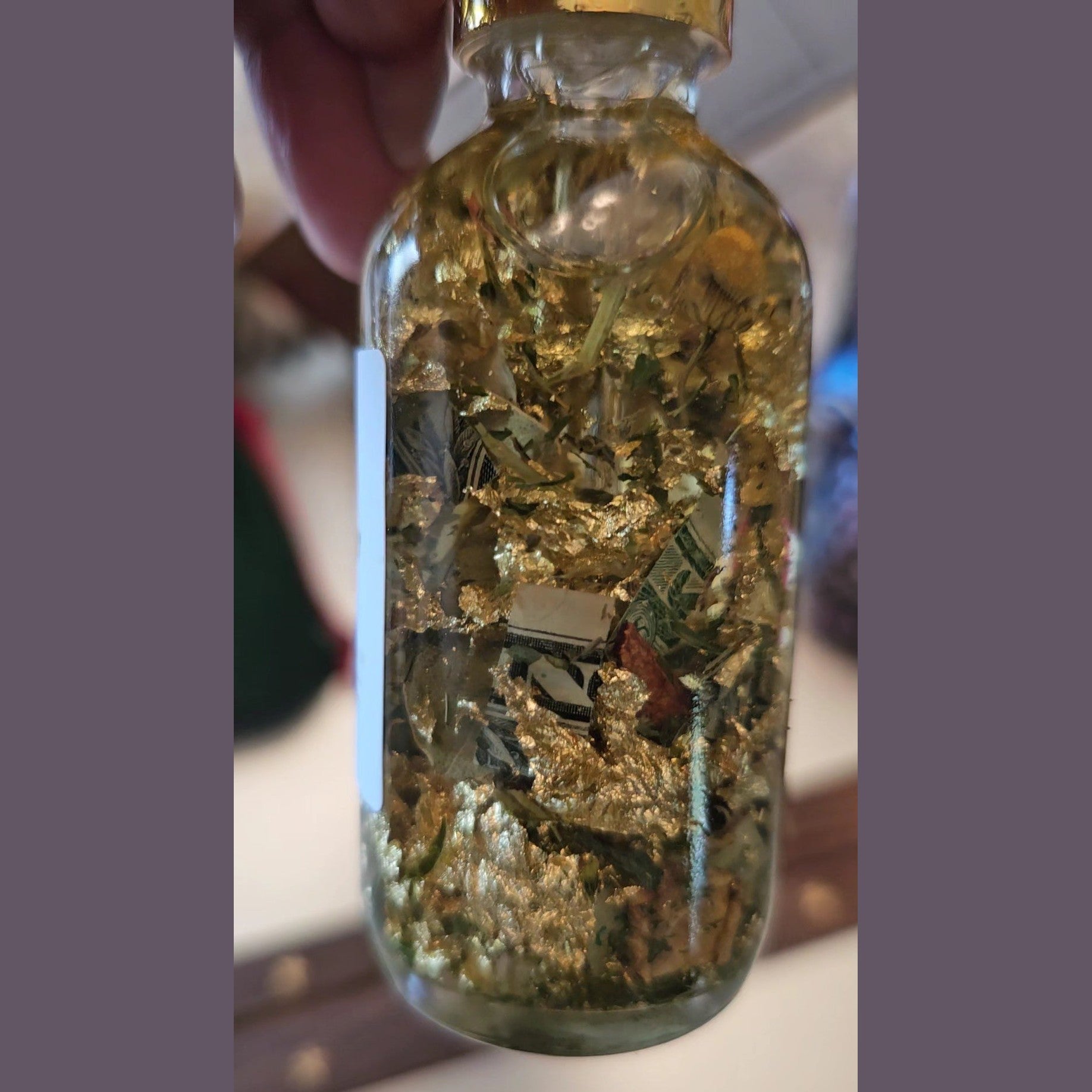 Glass bottle filled with gold leaf and herbs, held by a hand. Fast Money Oil - Luxury Hoodoo Conjure Oil for Quick Cash & Prosperity