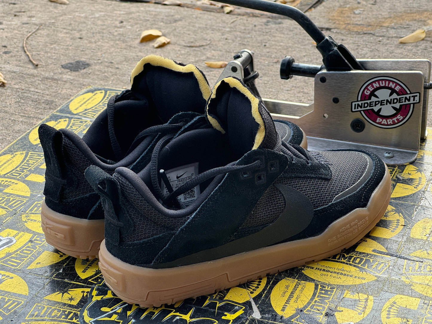 A pair of black Nike Air Max 2013 sneakers with yellow lining and gum soles, displayed next to a Genuine Parts hydraulic tool on a yellow and black floor mat. Nike SB Day One - Youth - FN4210-001