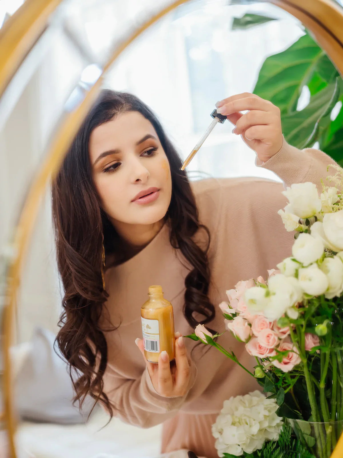 Luxury body oil with 24K gold shimmer in a glass dropper bottle, reflected in a mirror as a woman applies the oil while surrounded by white and blush flowers.