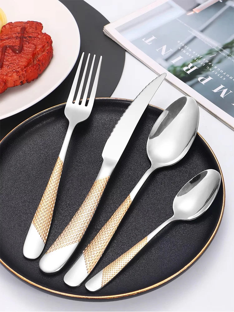 Set of silver cutlery with gold accents on a black plate, next to a plate of food and a magazine.