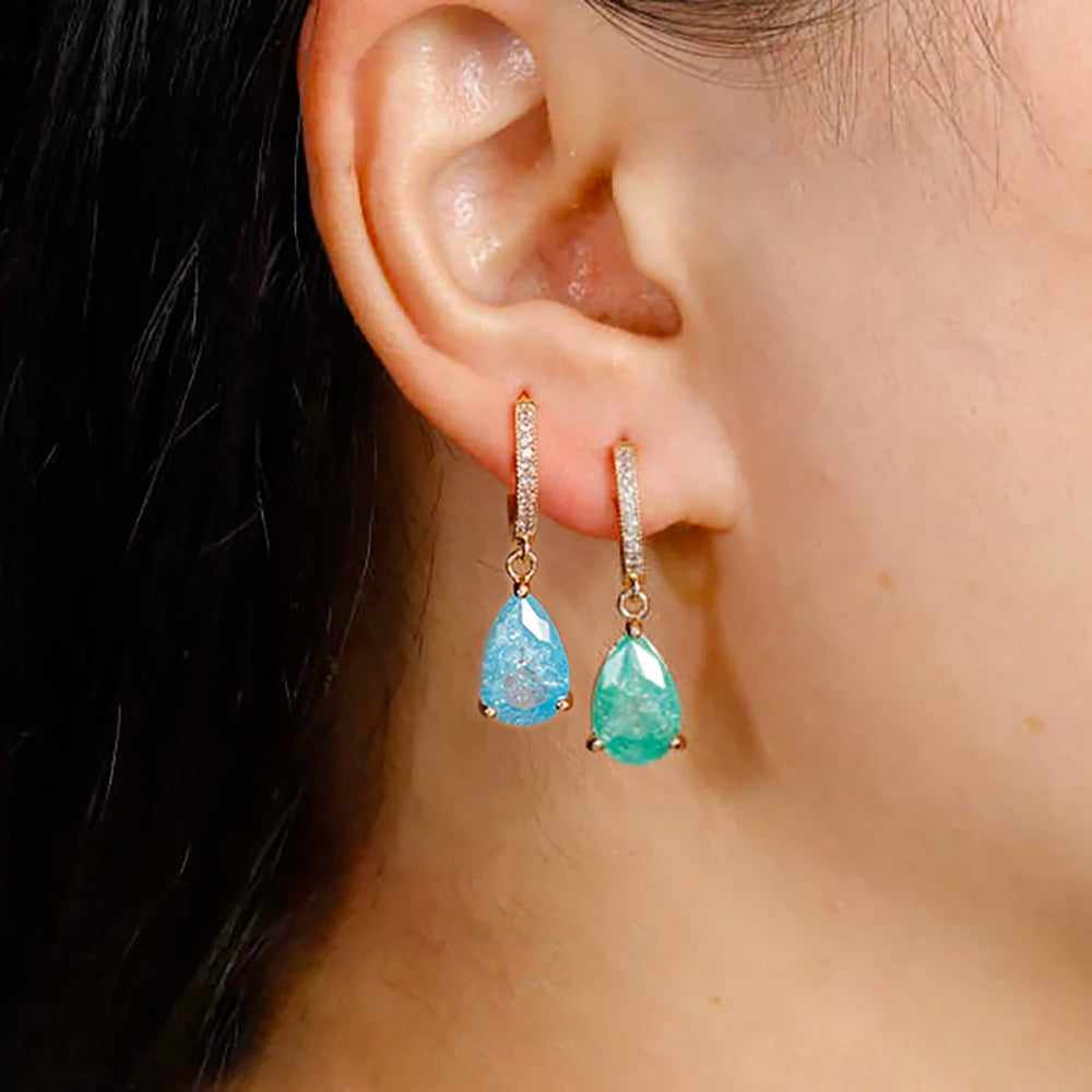 Close-up of a person wearing two teardrop-shaped earrings with blue and green gemstones.