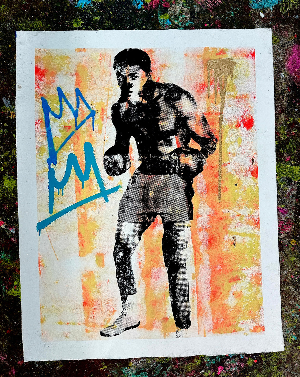 A square art print featuring a stylized black-and-white illustration of Muhammad Ali, set against a warm orange, yellow, and red abstract background with blue graffiti-style lettering, photographed on a textured dark surface. #2 Ali - Silk Screen, Acrylic & Spray Paint on canvas, 2024