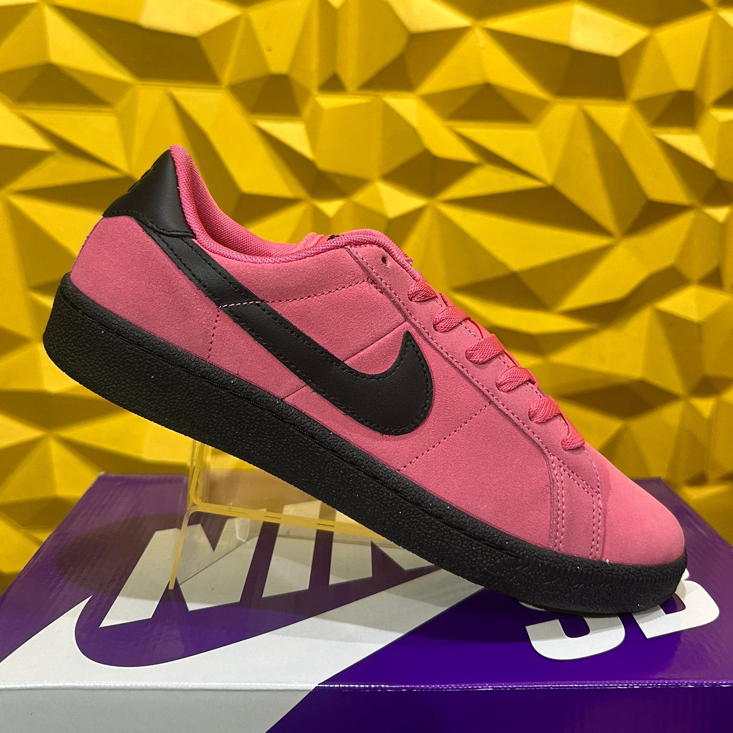 A pink Nike sneaker with a black Swoosh and black sole, displayed on a clear stand atop a purple Nike shoebox against a yellow geometric wall. Nike SB Zoom Tennis Classic FC