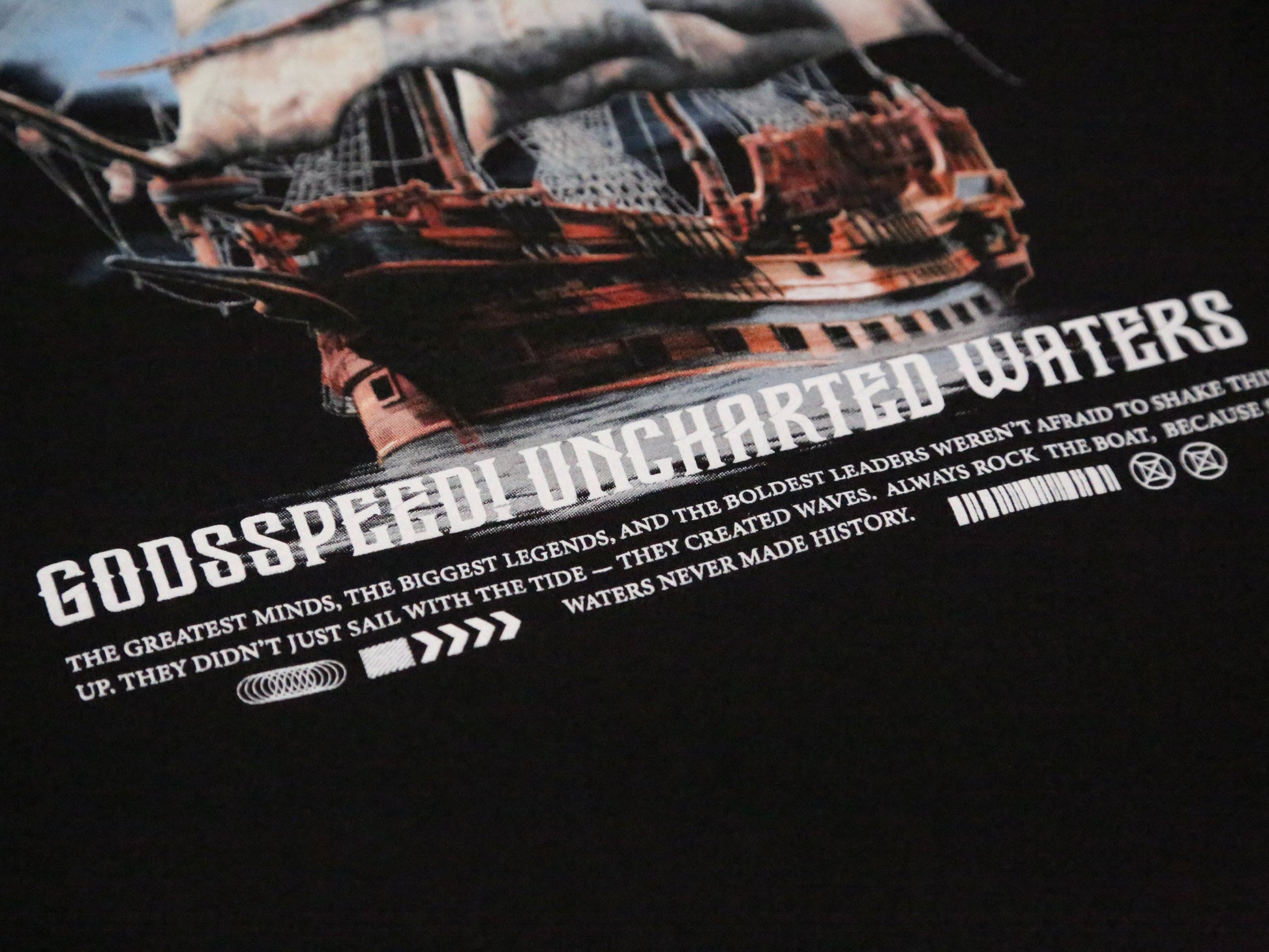 Close-up of a black graphic t-shirt featuring a distressed illustration of a sailing ship and bold white text reading 'GODSSPEED UNCHARTED WATERS' with smaller descriptive text below.