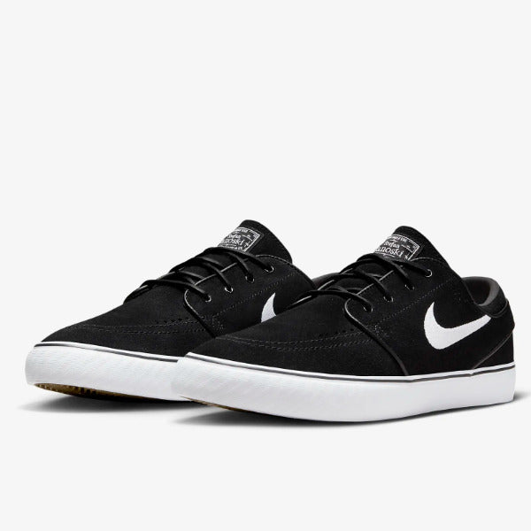 A pair of Nike low-top skate shoes in black canvas with a white Swoosh and white rubber sole, shown against a light gray background. Nike SB Janoski Black White