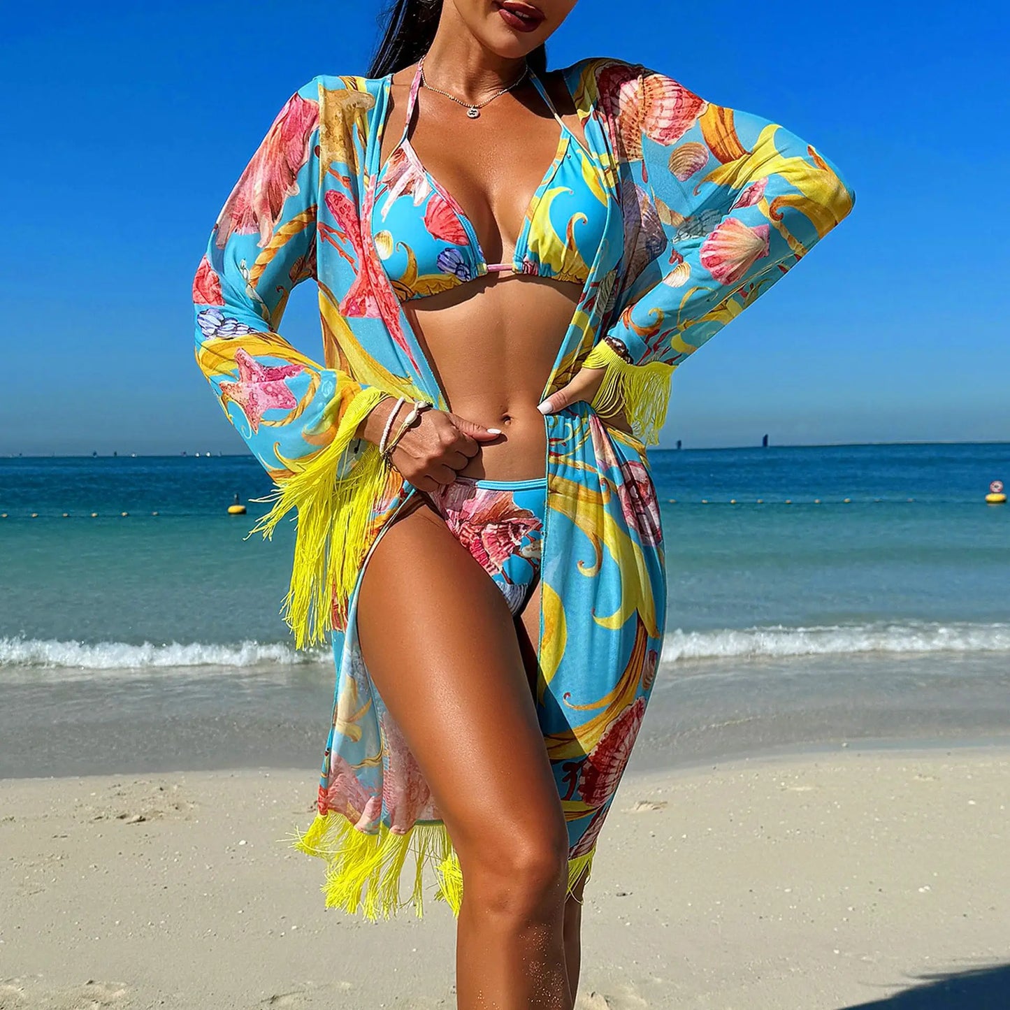 Woman in a colorful bikini and kimono standing on a beach with blue water and sky.