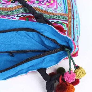 Handcrafted boho-style tote bag featuring a blue drawstring closure and vibrant, multi-colored pom-pom tassels, displayed against a clean white background.